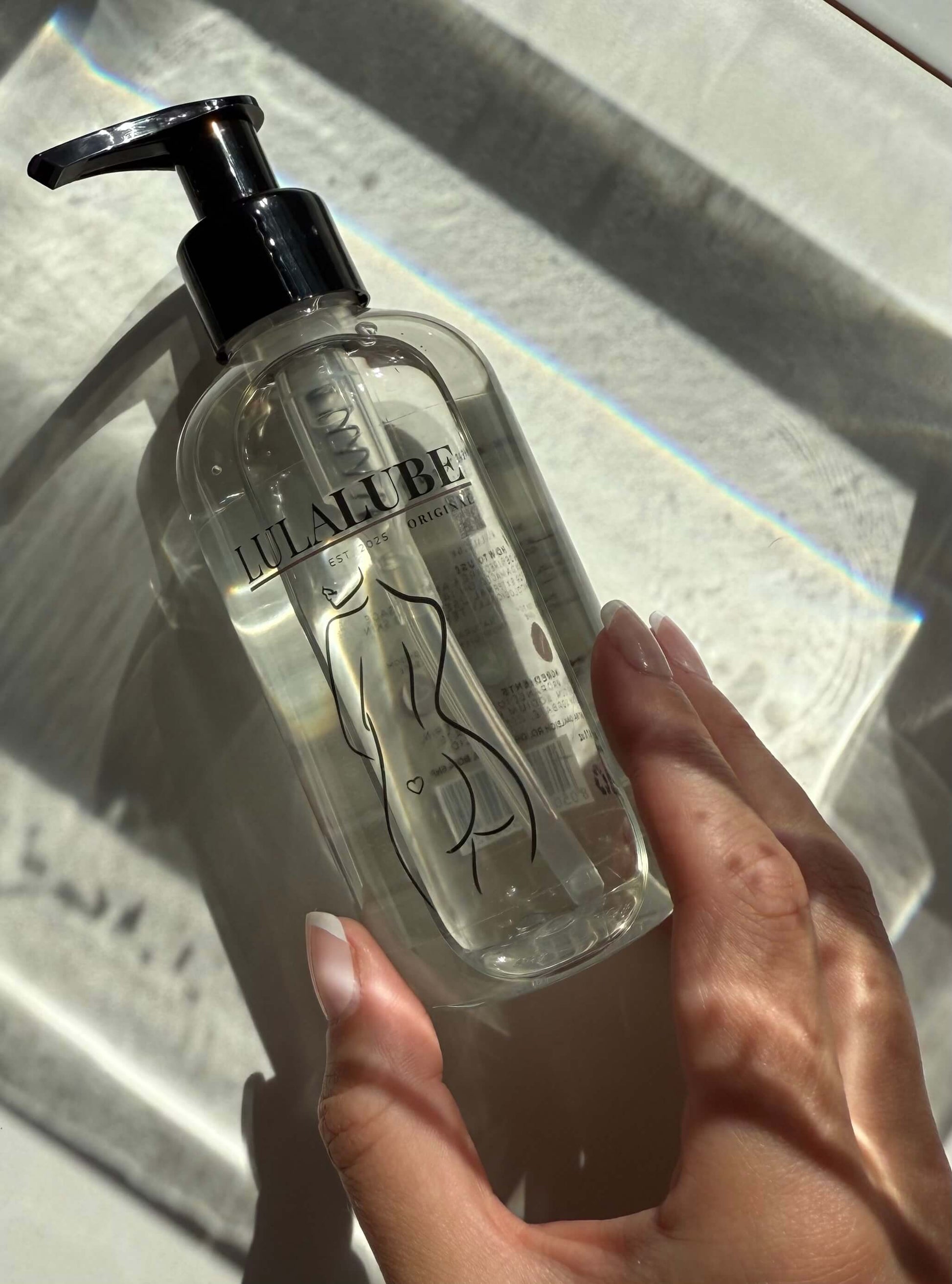 Hand holding a clear water-based lubricant with a pump against a reflective surface.
